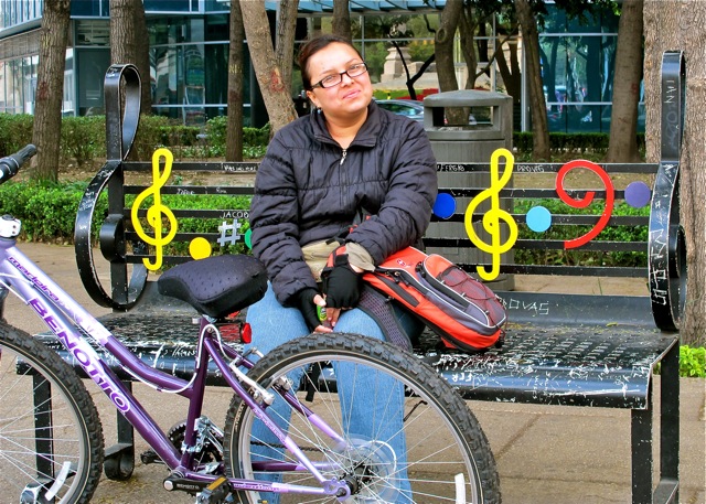 Smiling young woman sitting on a black iron bench with musical notations; her lavender bike in front.